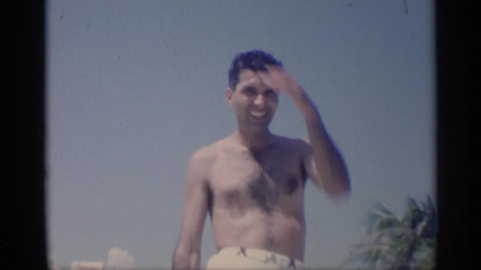 1959 California 8mm frame showing a shirtless adult man smiling toward the camera while touching his hair, with palm foliage and bright sky behind him.