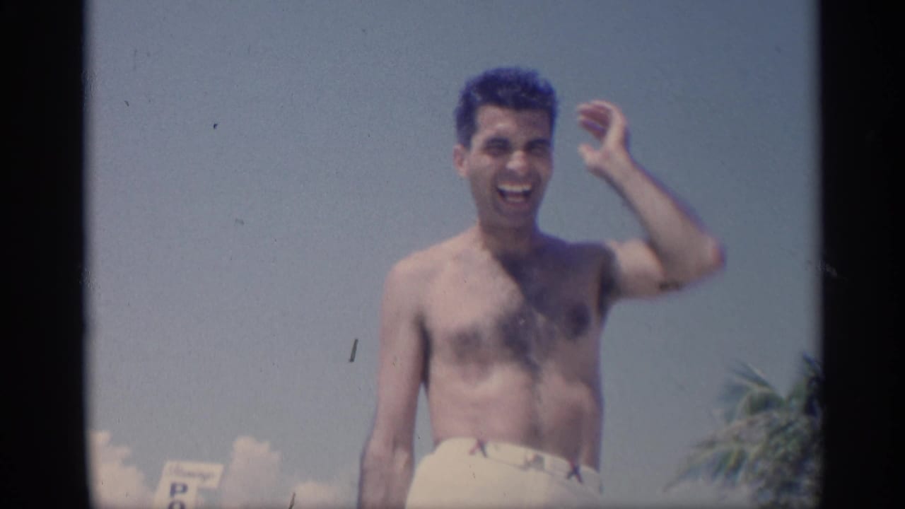 Shirtless young man in white swim trunks smiles and raises one hand on a sunny beach.