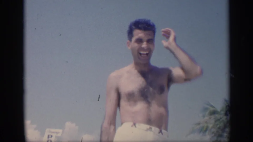 Young man in white swim trunks smiles toward the camera on a 1959 California beach, with blue sky and palm trees behind him.