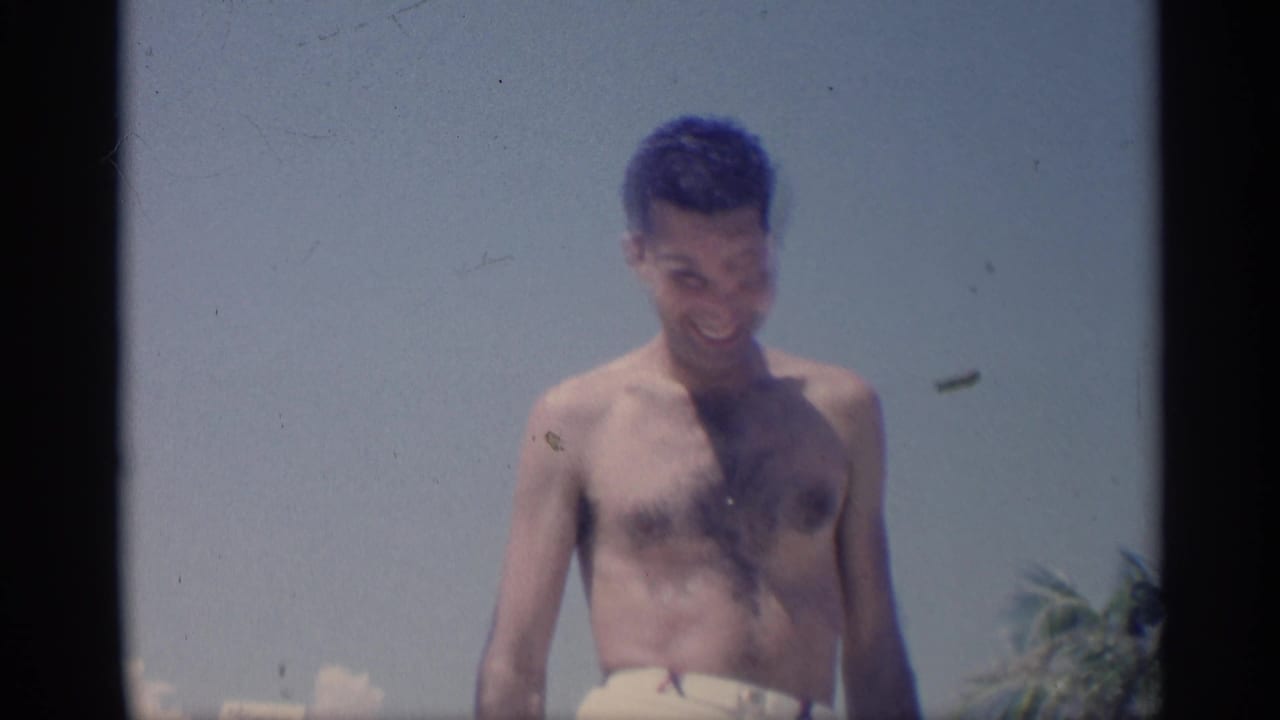 Shirtless young man looks down while standing on the beach with palms in the background.