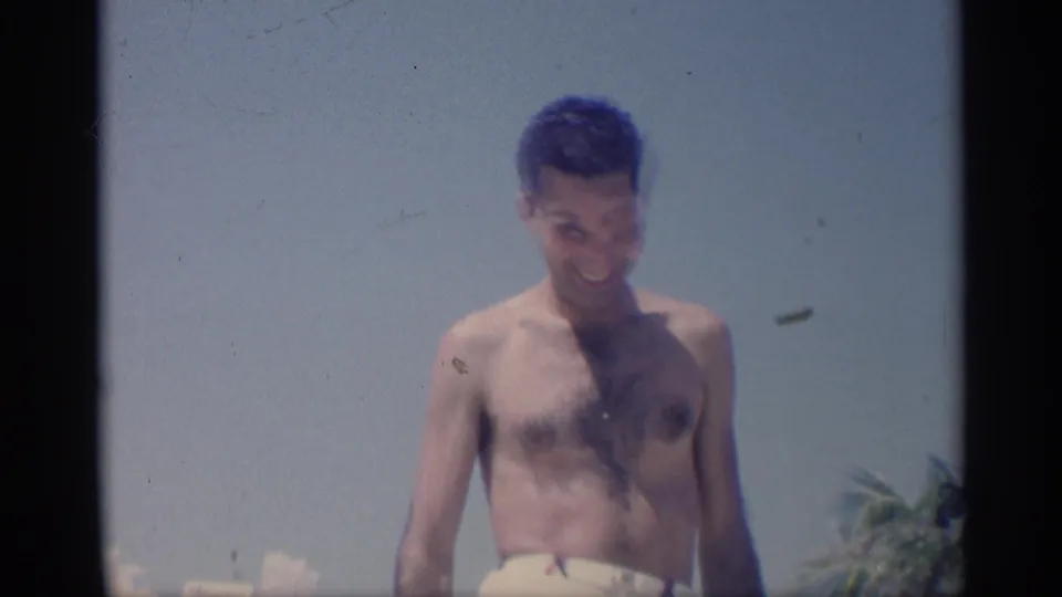 The young man stands shirtless in white swim trunks on a California beach, looking downward during the 1959 home-movie shot.