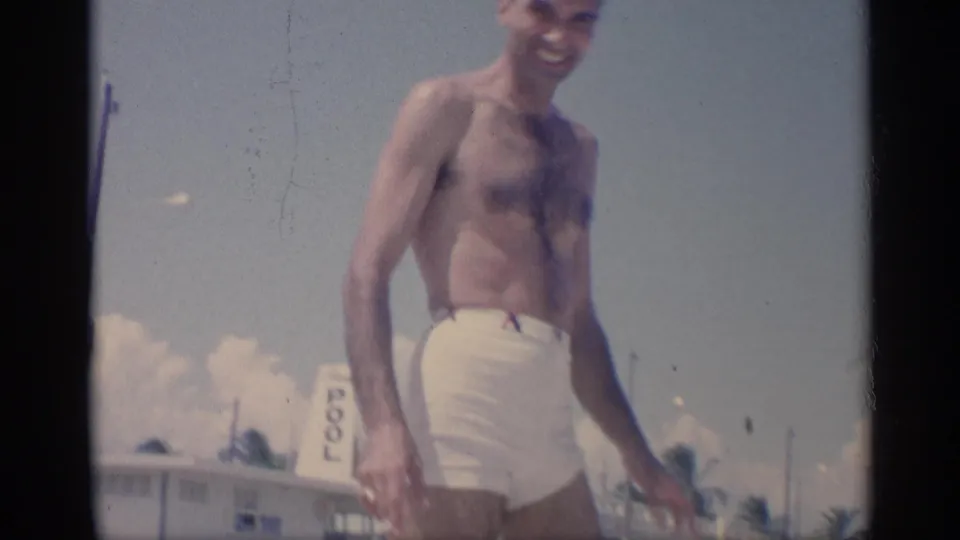 A shirtless young man in white trunks turns near a California beach setting, with a visible pool sign and palms in the background.
