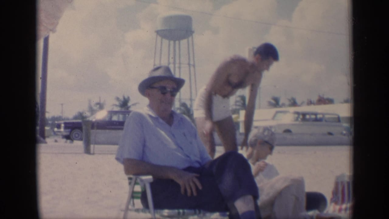 1959 California beach frame with a seated man in a hat, another adult bending nearby, classic cars, and a water tower in the background.