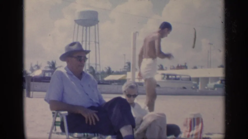 1959 California beach view with a seated man in sunglasses, a man walking in white swim trunks, parked cars, and a water tower.