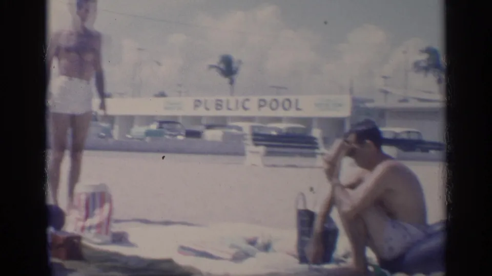 1959 California beach frame showing adults near towels and chairs, a public pool sign, classic cars, palm trees, and bright sand.