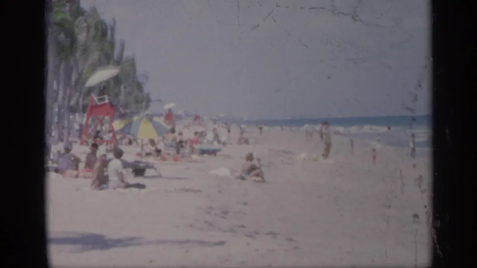 Wide 1959 California beach view with scattered visitors on sand, palm trees, umbrellas, a lifeguard-style structure, swimmers, and ocean water.
