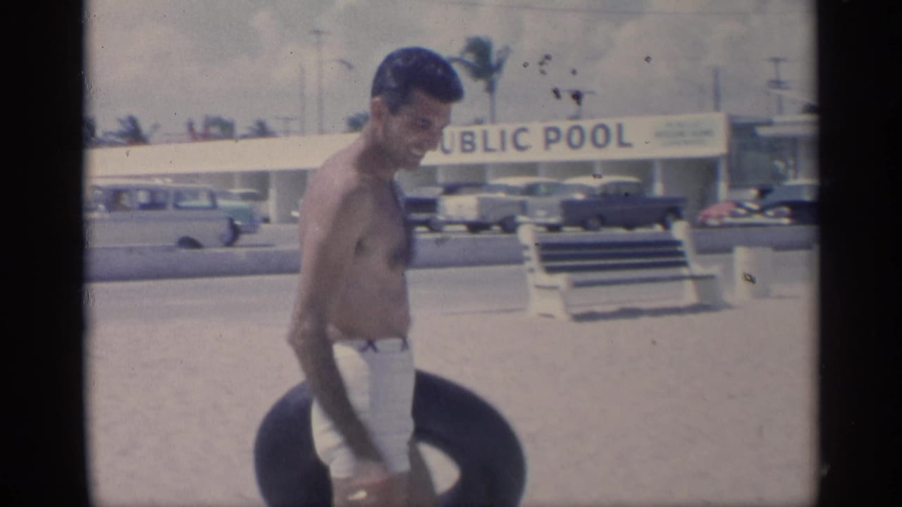 Shirtless man in white swim trunks walks with a black inner tube near a public pool sign.