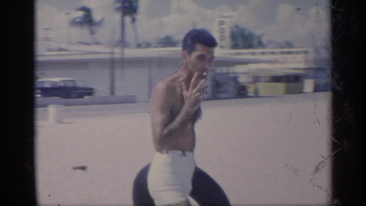 Man with inner tube holds a cigarette while standing on a sandy beach area.