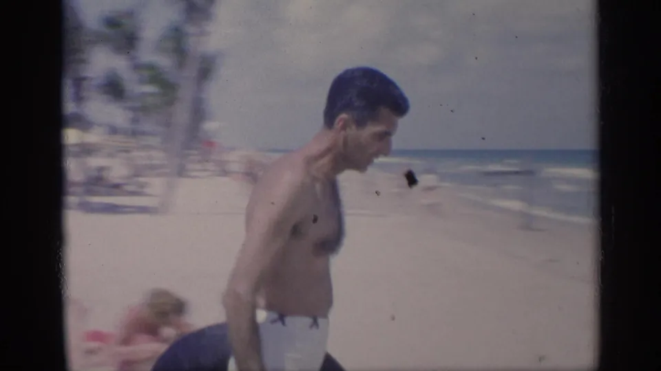 A man in white swim trunks carries a black inner tube along a sunlit California beach, with surf and beachgoers in the background.