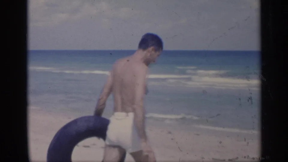 The man walks away toward the ocean with a black inner tube, framed by pale sand, blue water, and the horizon line.