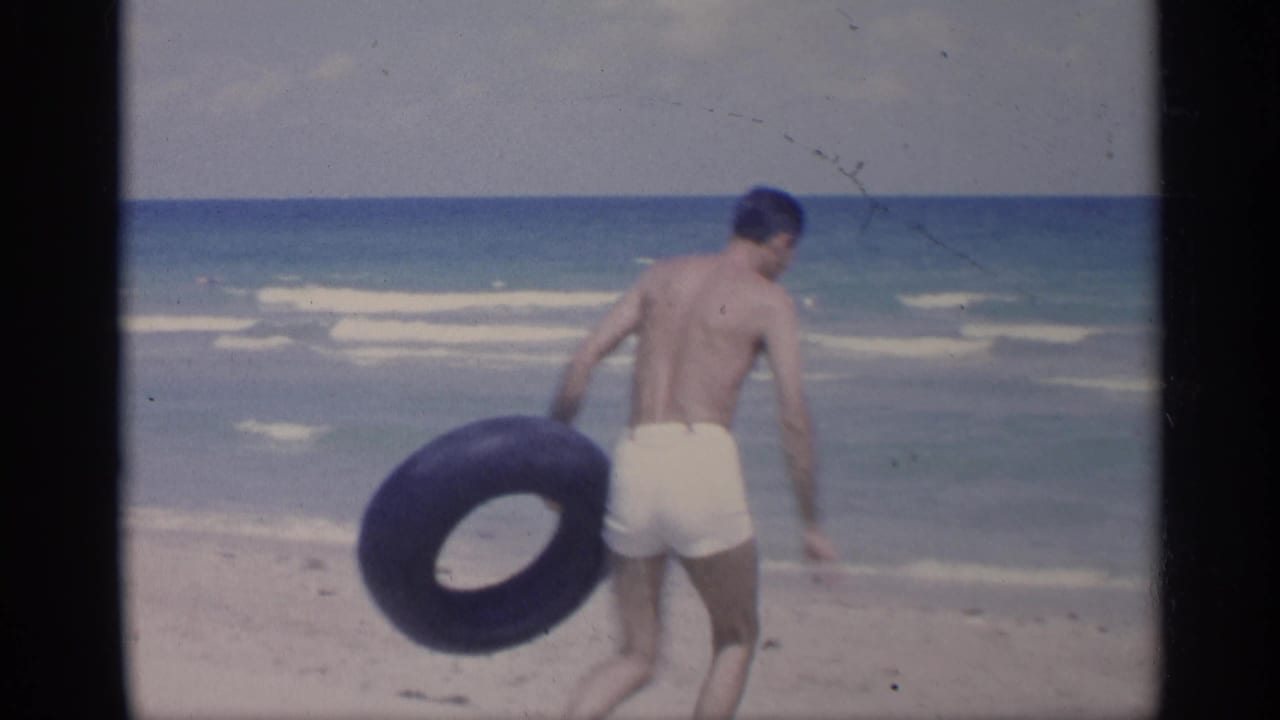 Man in white swim trunks walks on a sandy beach carrying a black inner tube.