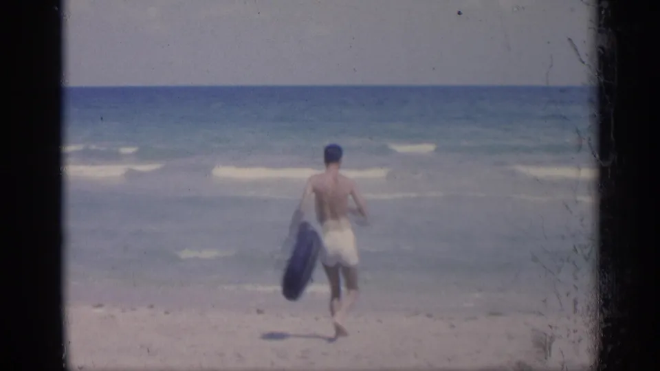 The man moves down the beach toward the surf with a black inner tube under a bright coastal sky.