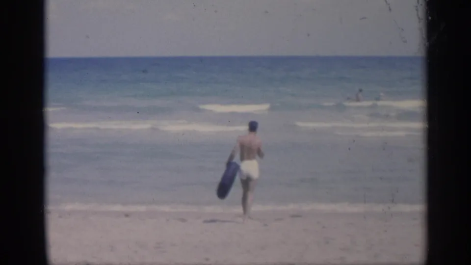 From farther back, the 8mm frame shows the man nearing the waterline with distant swimmers offshore.