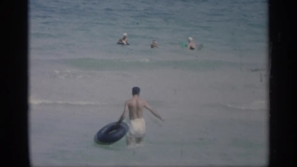 The man reaches shallow ocean water with his inner tube while several swimmers are visible beyond the small waves.