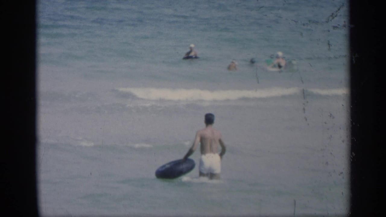 Young man in white swim trunks carries a black inner tube into shallow surf.