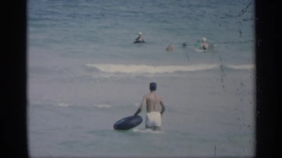 1959 California beach footage showing a young man in white trunks wading into the ocean with a black inner tube as swimmers float offshore.