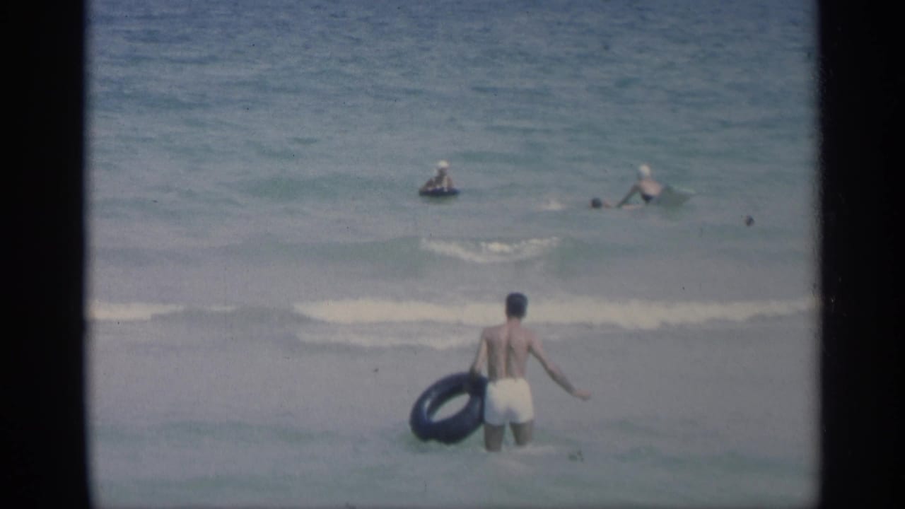 Man stands in shallow waves holding a black inner tube as swimmers are offshore.
