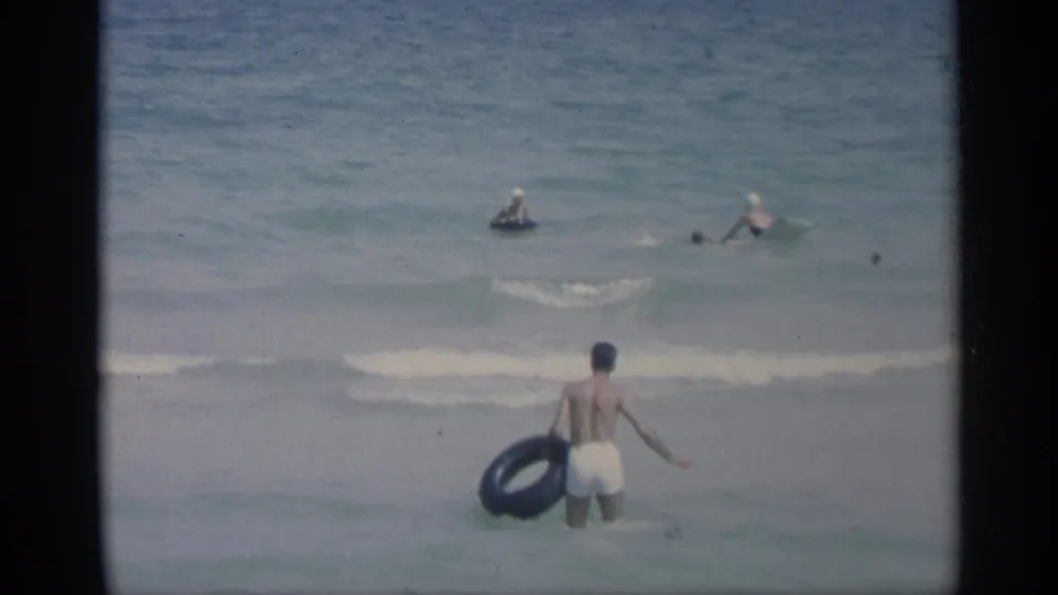 A man in white trunks holds a black inner tube near the shoreline in California surf, with several swimmers visible in the water beyond.