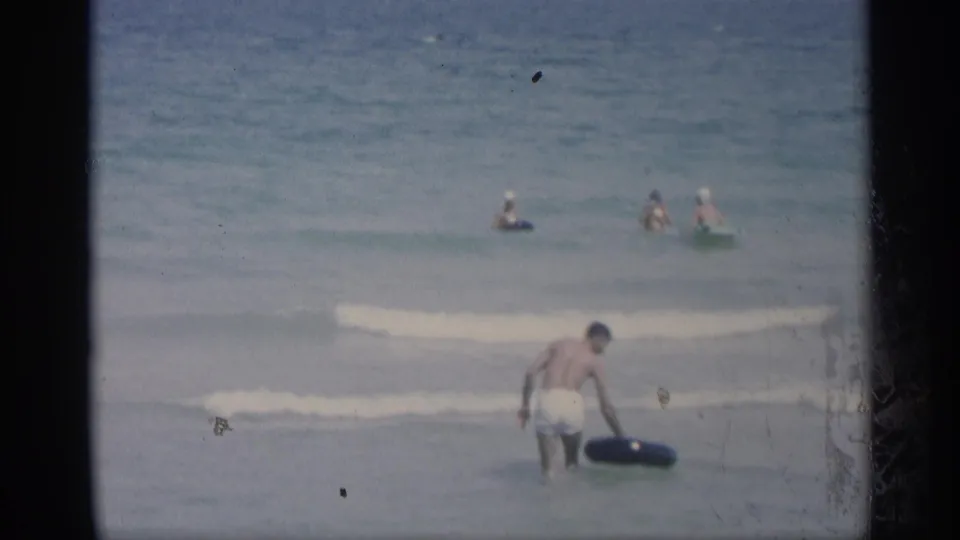 The swimmer bends toward a black inner tube in shallow California surf while small groups of bathers remain farther out in the ocean.