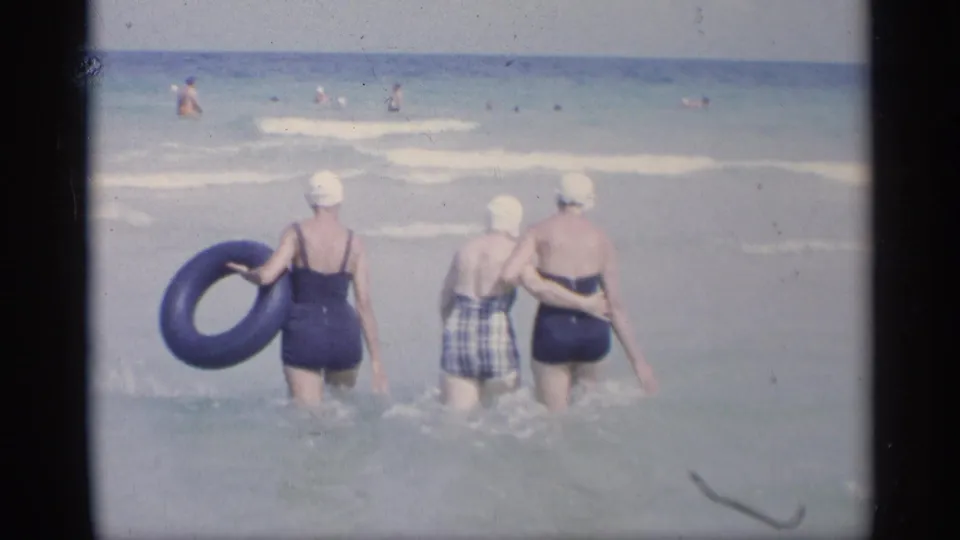 Three women in vintage one-piece swimsuits and swim caps wade into the California surf, with one holding a black inner tube.