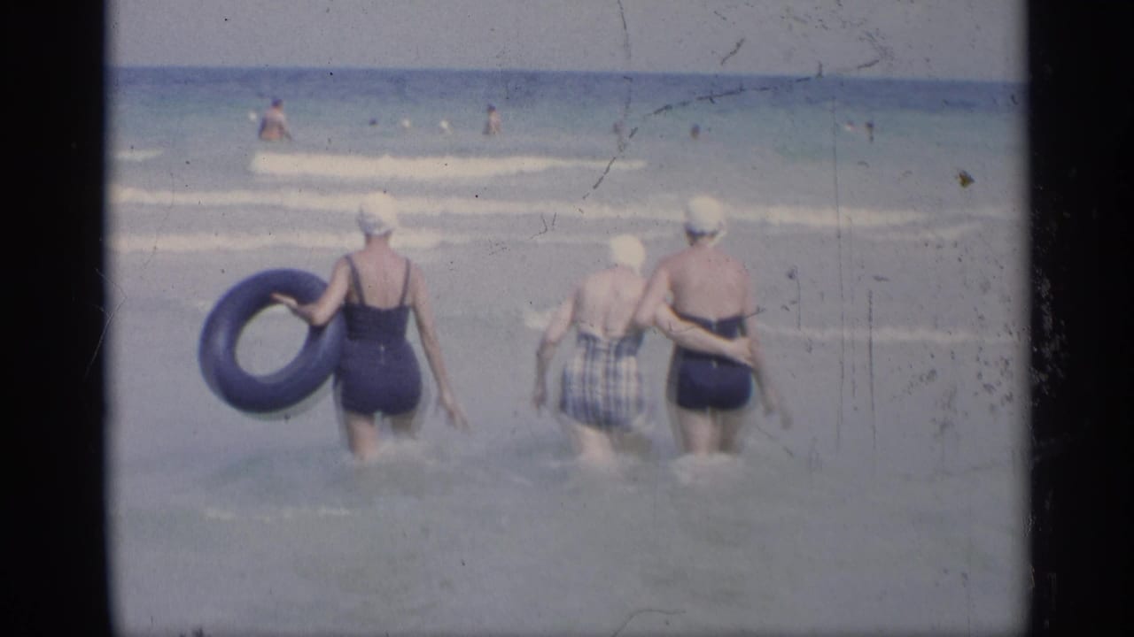 Three women in swim caps wade into the ocean, one carrying a blue inner tube.