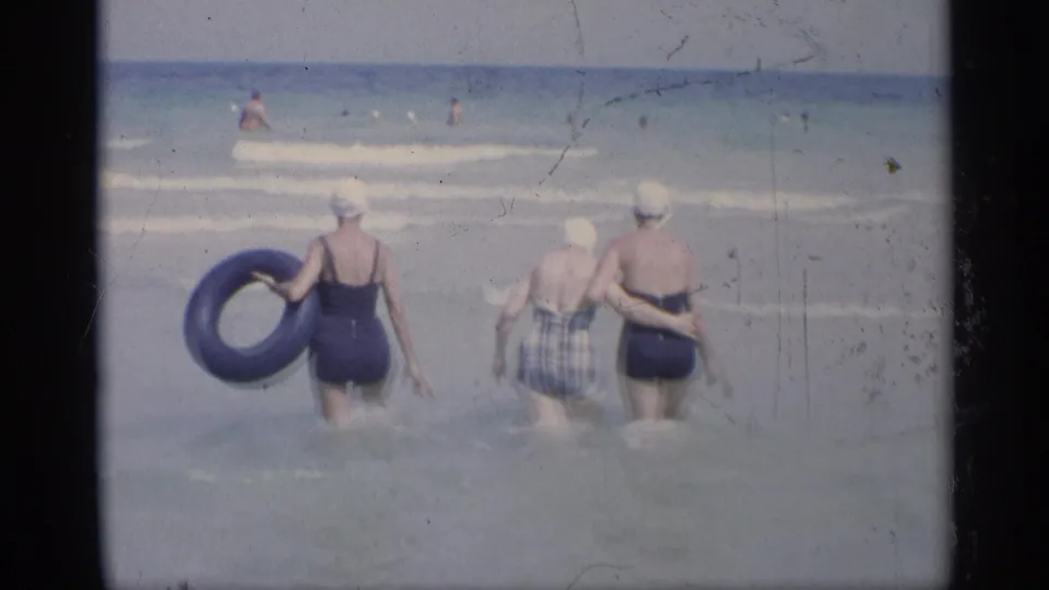 1959 California 8mm frame of three women entering shallow surf, with one holding a blue inner tube and distant bathers beyond.