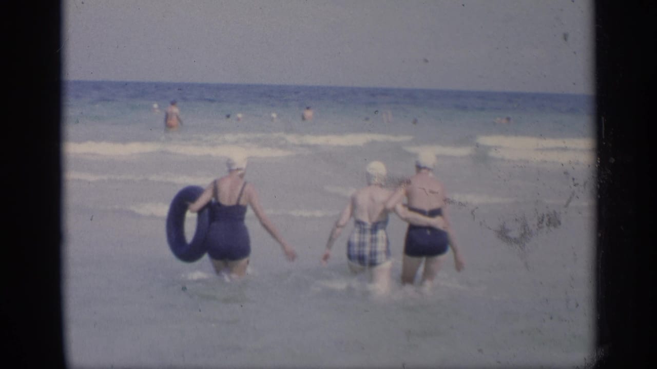 Three women continue walking through shallow surf with distant swimmers offshore.