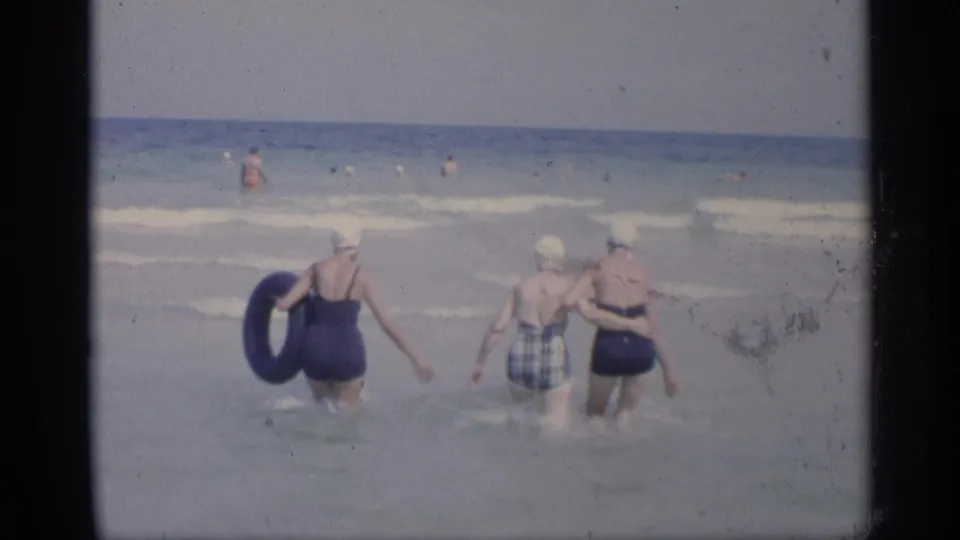 Three women in mid-century swimsuits and caps wade farther into the California ocean, one carrying a blue inner tube.