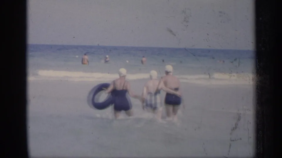 Soft 8mm view of three women in swim caps pausing in the surf, with the blue inner tube prominent on the left.