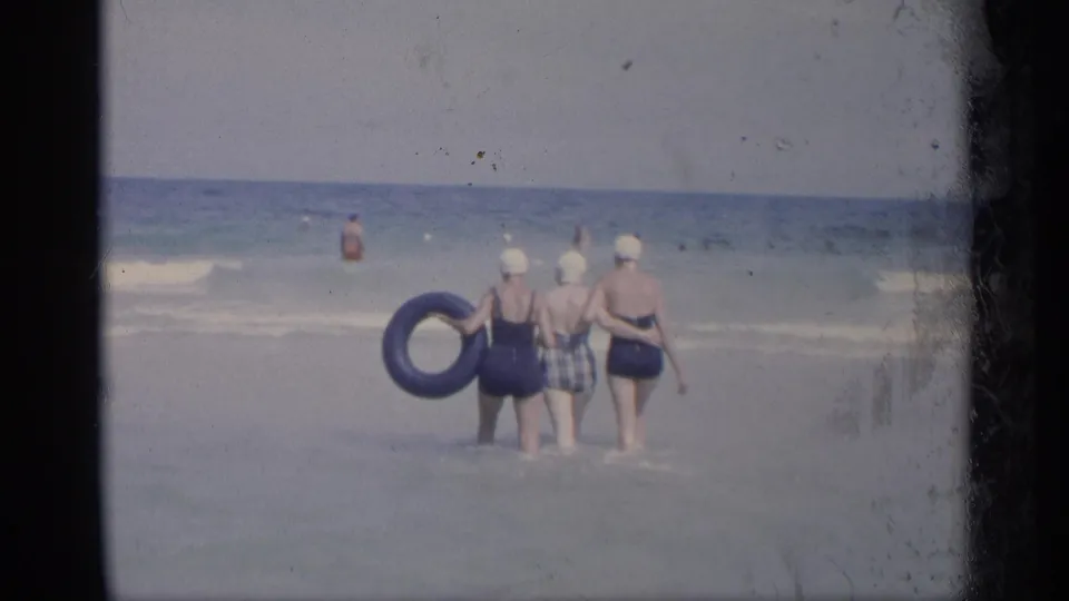1959 California beach frame showing three women from behind in shallow surf, with a blue inner tube and distant swimmers offshore.