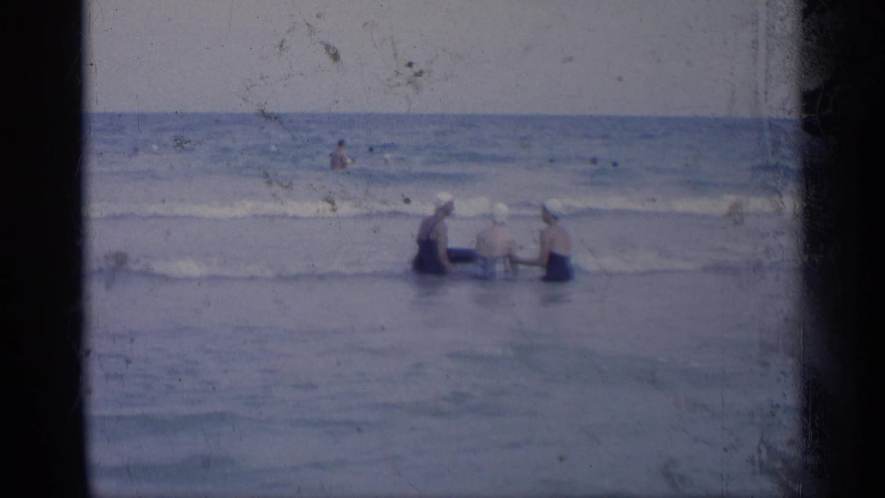 In a 1959 California beach home-movie frame, three women in swim caps wade into shallow surf while holding a blue inner tube.