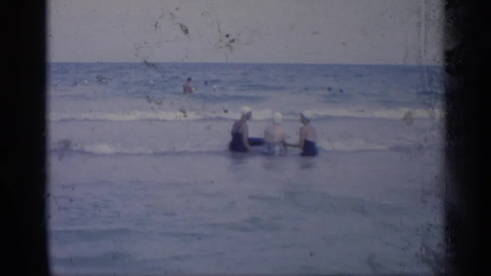 In a 1959 California beach home-movie frame, three women in swim caps wade into shallow surf while holding a blue inner tube.