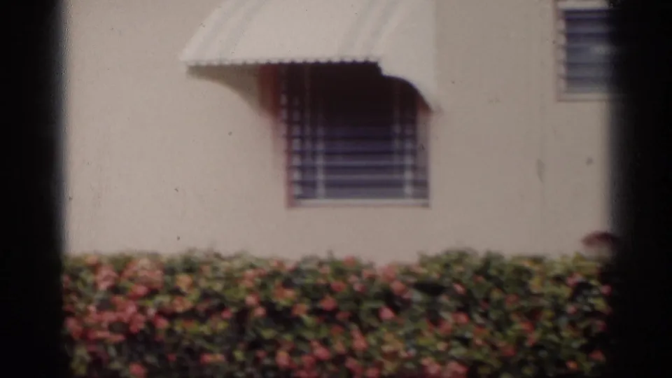 A brief 1959 California home-movie view shows a pale residential-style building exterior with an awning, window bars, shrubs, and lawn.