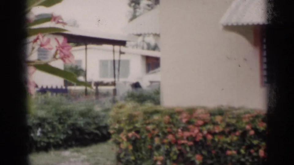 A handheld 1959 California frame looks across landscaped grounds between pale buildings, with flowers, awnings, shrubs, and a patch of lawn.