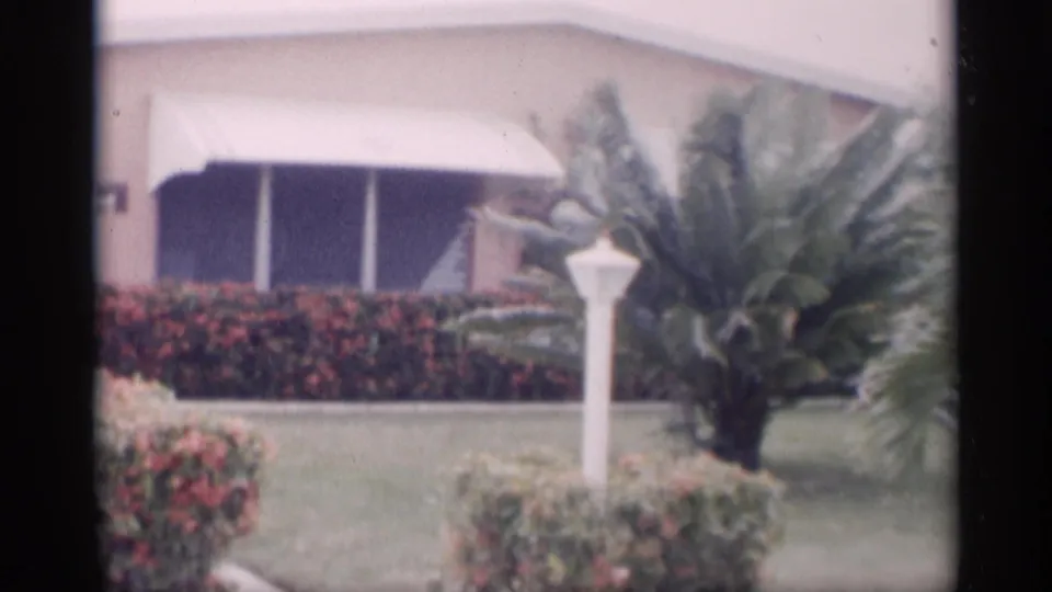 1959 California 8mm frame showing a manicured yard with a white lamppost, red flowering hedge, palm plant, lawn, and house awning.