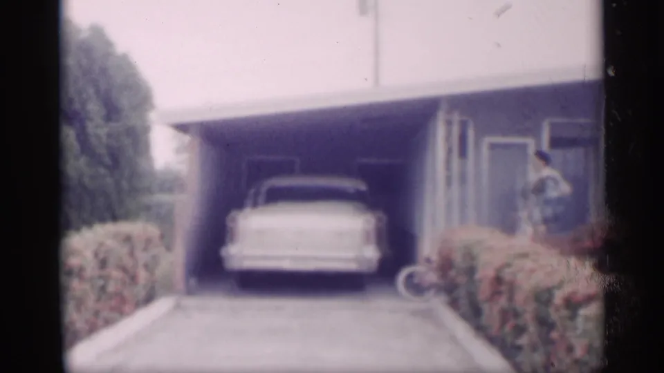 1959 California suburban driveway with a white classic car under a carport, hedges, bicycle, house exterior, and a distant person near the entrance.