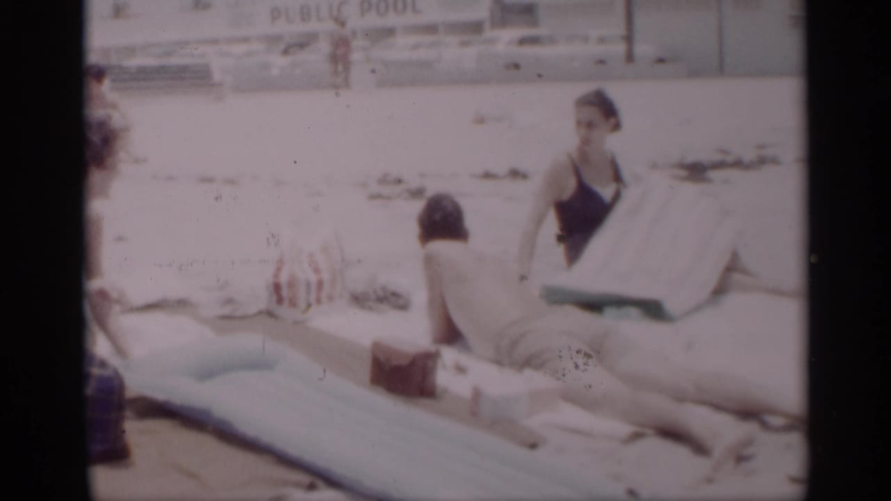 Man and woman resting on a sandy beach near towels and an inflatable mattress.