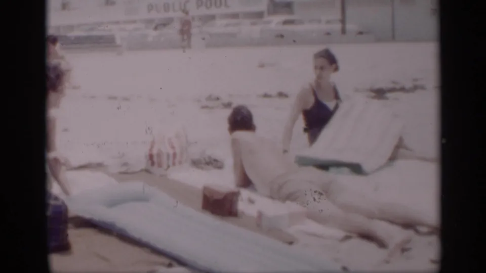 1959 California beach home-movie frame showing a woman seated on a towel and a man lying nearby, with a public pool sign in the background.