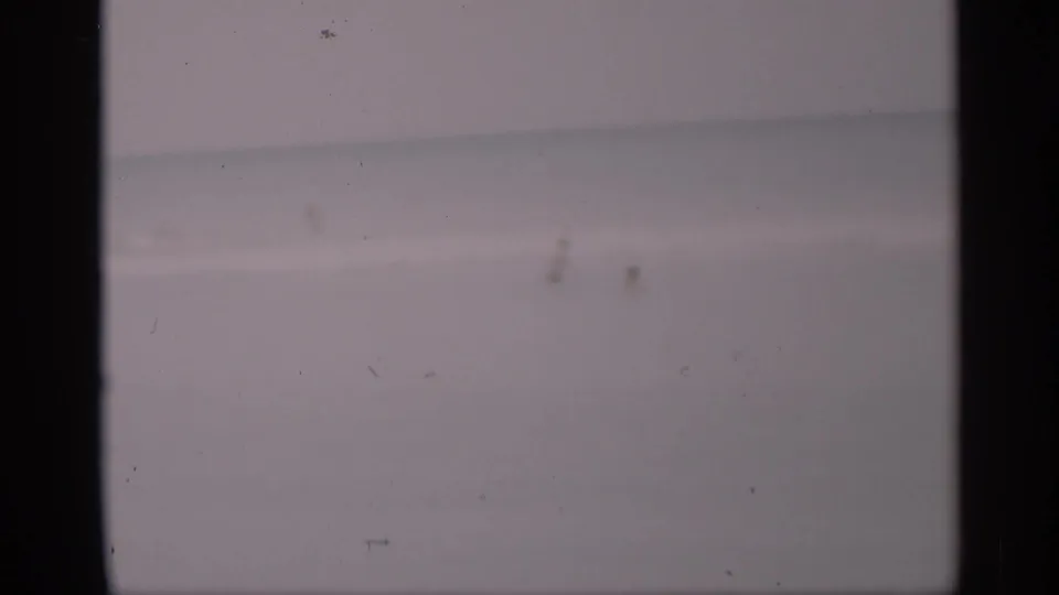 Faded 8mm frame of distant people near the California surf, with minimal shoreline detail and a broad pale ocean background.