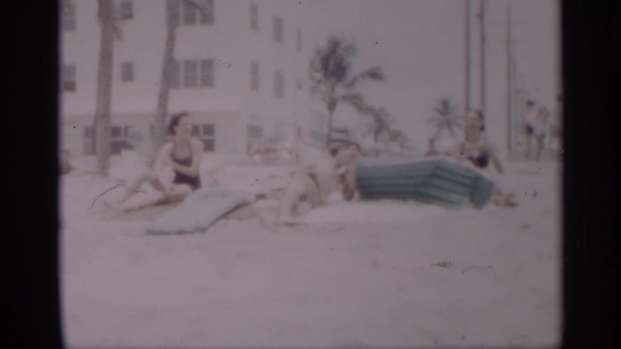 Adults relaxing on sand near a green inflatable mattress with palm trees and a white building behind them.