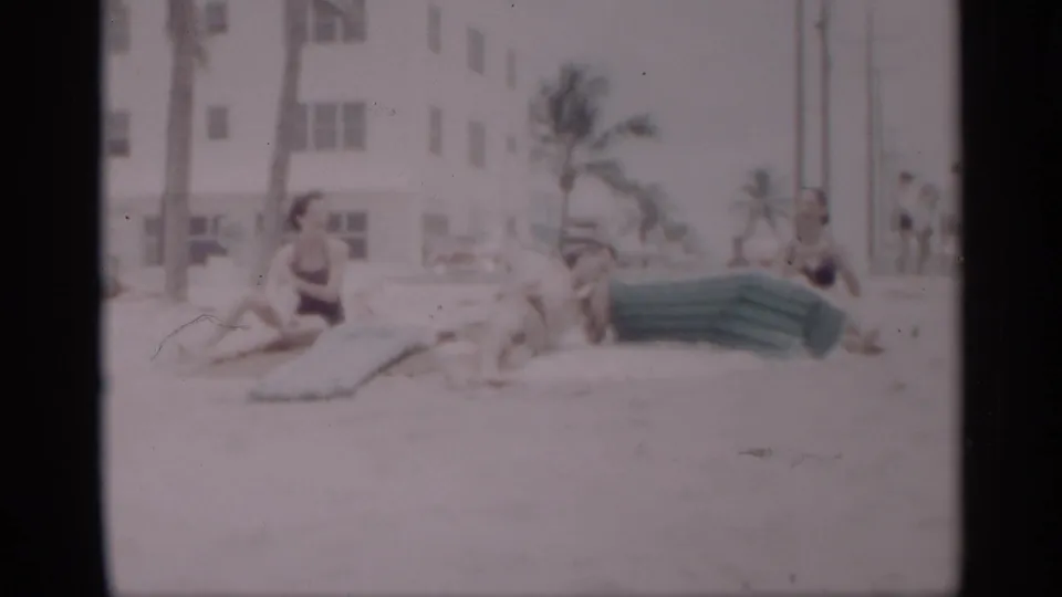 1959 California beach home-movie frame showing adults in swimsuits lounging on sand beside a green inflatable mattress near palms and a white building.