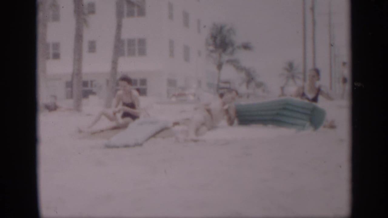 Beachgoers sit and recline on sand beside a green inflatable mattress in front of palms and a white building.