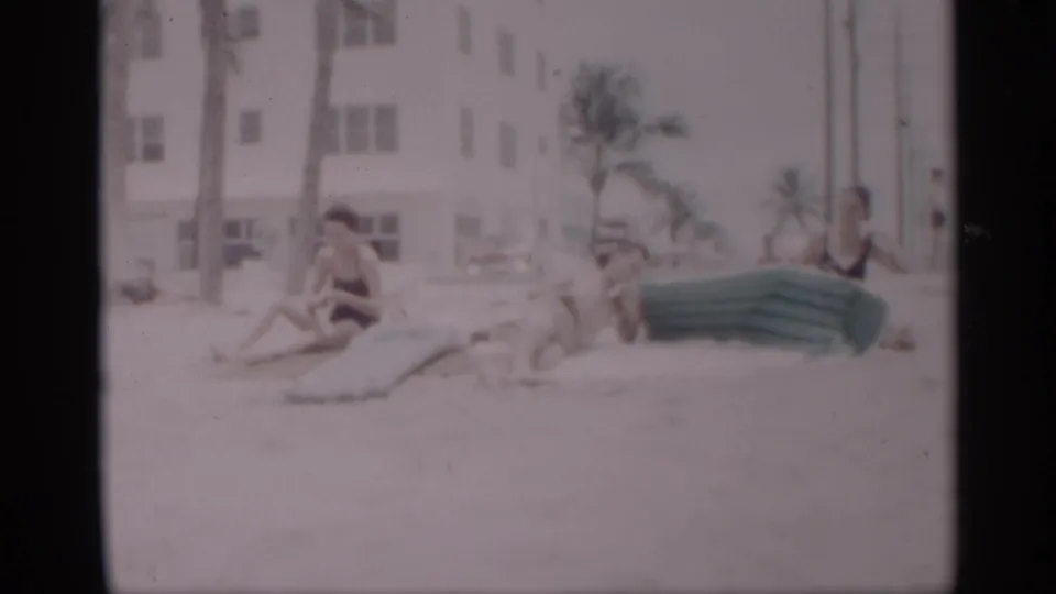 1959 California beach scene with adults seated and reclining on sand near a green inflatable mattress, palm trees, and a white building.