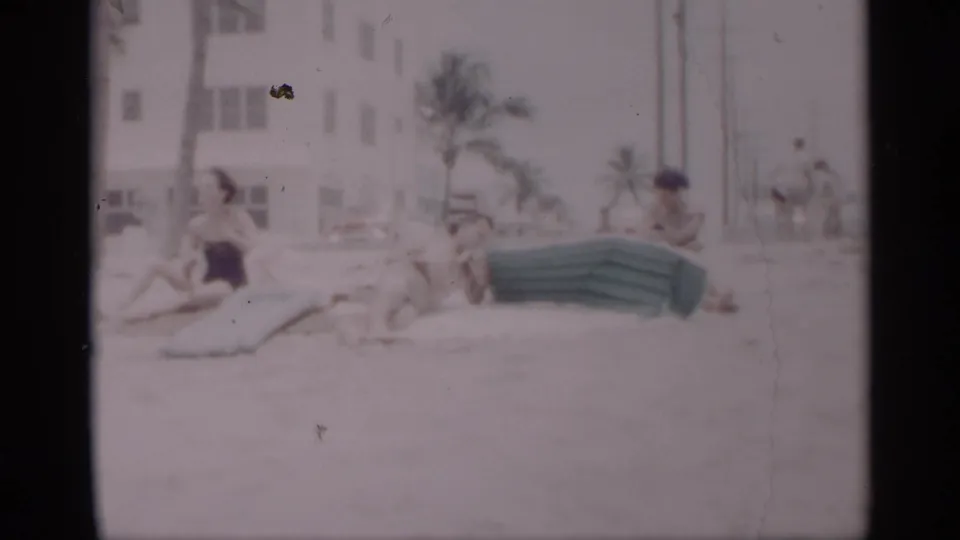 Soft 8mm frame of 1959 California beachgoers relaxing near a green inflatable mattress, with palm trees and a white building behind them.