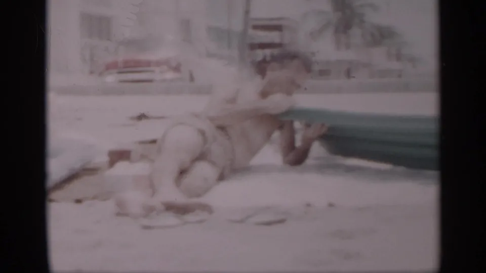 1959 California beach home-movie frame showing a man kneeling near a green inflatable mattress, with towels or small items on the sand.
