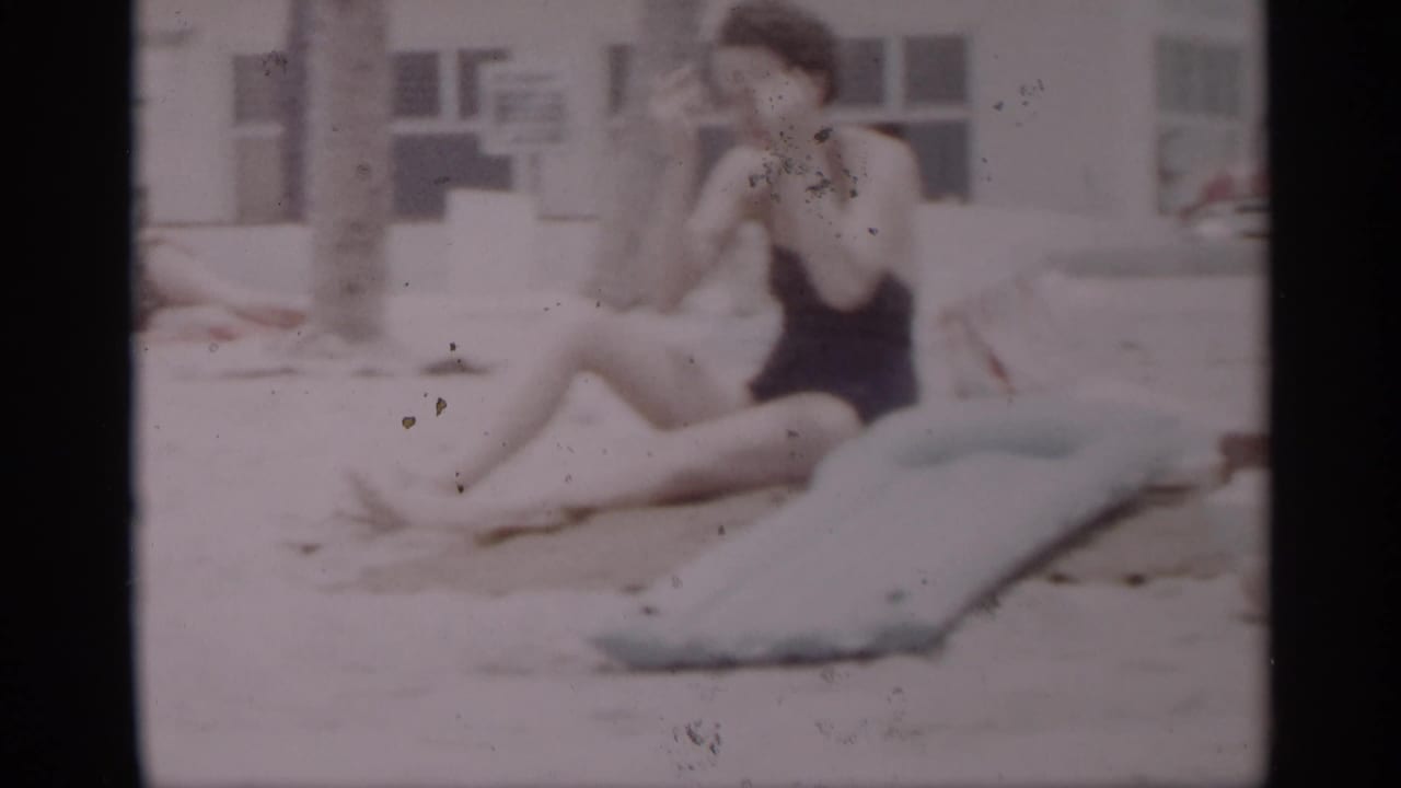 Woman in a dark one-piece swimsuit sits on sand near a light blue inflatable mattress.