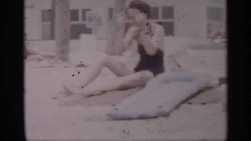 1959 California beach frame showing a woman seated on sand beside a light blue inflatable mattress, with a building and beach area behind her.
