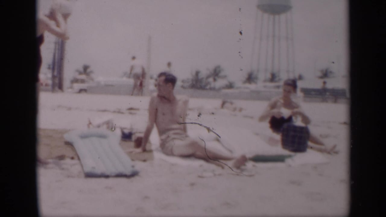 Shirtless man sits on a towel while a woman sits nearby on a sunny beach.