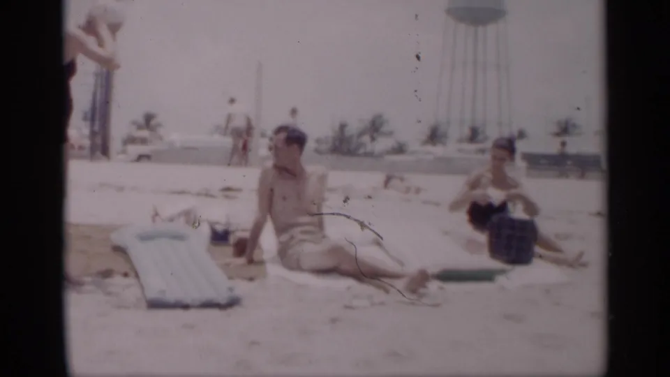 1959 California beach scene with a shirtless man seated on a towel, a woman nearby, an inflatable mattress, palm trees, and a water tower in the background.