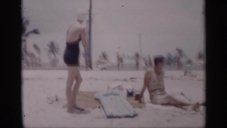1959 California beach frame showing a woman standing near a seated shirtless man, with an inflatable mattress, palm trees, and distant beach activity visible.